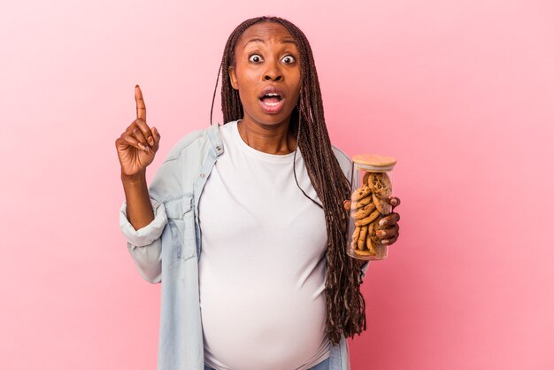 Jonge Afro-Amerikaanse zwangere vrouw met cookies geïsoleerd op roze achtergrond met een idee, inspiratie concept.