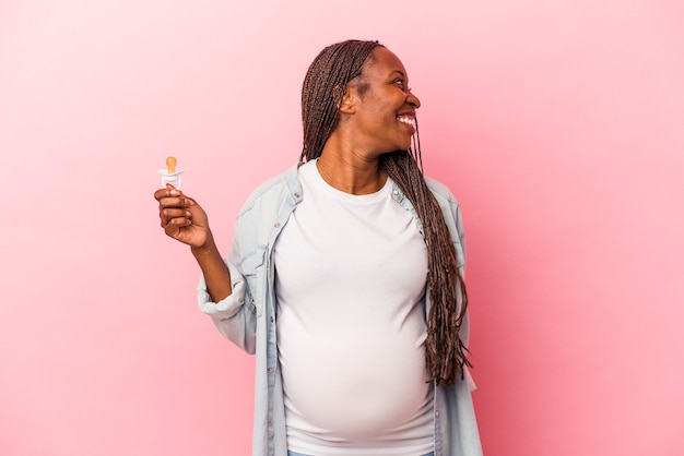 Jonge Afro-Amerikaanse zwangere vrouw met fopspeen geïsoleerd op roze achtergrond kijkt opzij glimlachend, vrolijk en aangenaam.