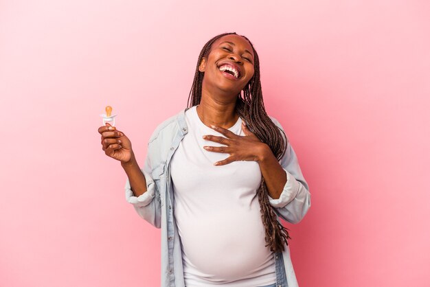 Jonge Afro-Amerikaanse zwangere vrouw met fopspeen geïsoleerd op roze achtergrond lacht hardop terwijl ze de hand op de borst houdt.