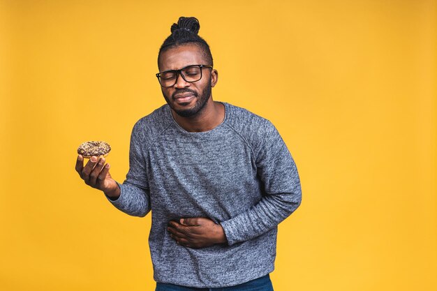 Jonge afro-amerikaanse zwarte man met afro-haar met donut geïsoleerd over gele achtergrond met de hand op de maag, pijnlijke ziekte die zich onwel voelt. Pijn concept.