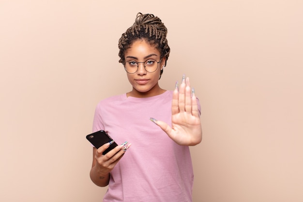Jonge afro-vrouw die ernstig, streng, ontevreden en boos kijkt die open palm toont die stopgebaar maakt. slimme telefoon concept