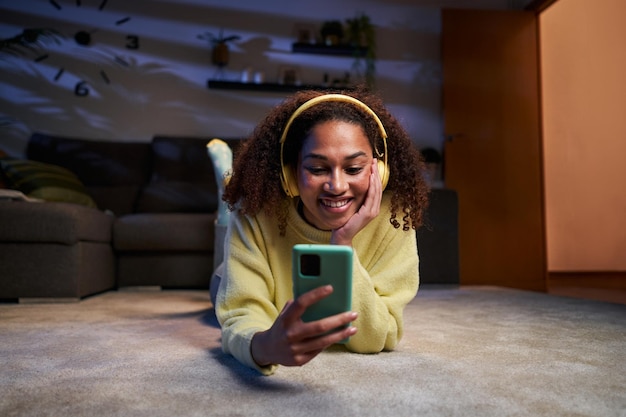 Jonge afro vrouw glimlacht naar mobiele telefoon met koptelefoon op liggend tapijt's nachts thuis