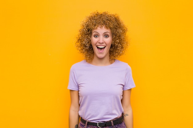 Jonge afro vrouw op zoek blij en aangenaam verrast, opgewonden met een gefascineerde en geschokte uitdrukking tegen oranje muur