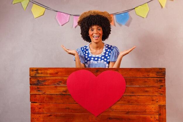 Jonge afrovrouw gekleed voor junina-feest achter een houten plank met een hart achter een houten plank met een hart