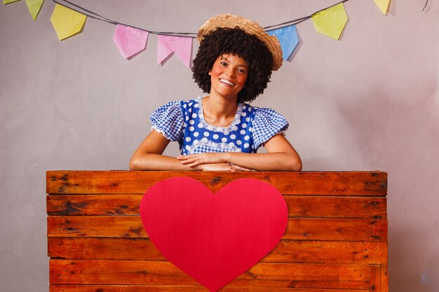 Jonge afrovrouw gekleed voor junina-feest achter een houten plank met een hart