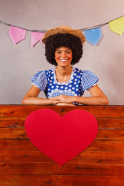 Jonge afrovrouw gekleed voor junina-feest achter een houten plank met een hart