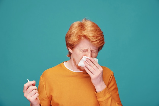 Jonge allergische man die papieren zakdoek bij zijn neus houdt terwijl hij niest en anti-allergiespray gebruikt voor camera geïsoleerd