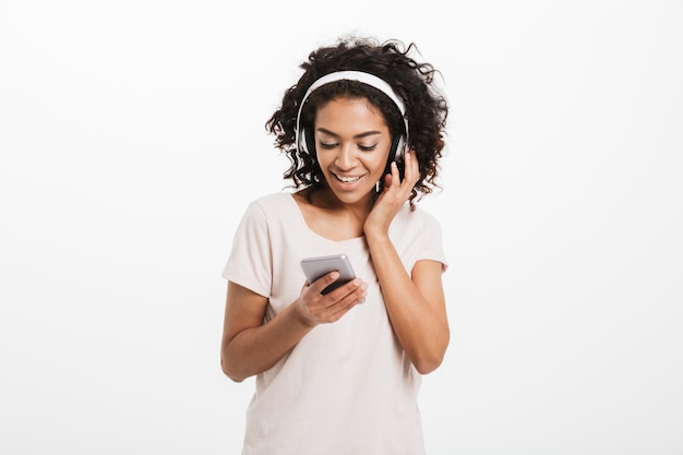 Jonge Amerikaanse vrouw met krullend kapsel dat aan muziek luistert en van favoriet lied via draadloze oortelefoons en smartphone geniet, die over witte muur wordt geïsoleerd