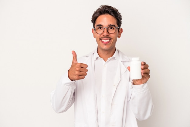 Jonge apotheker gemengd ras man met pillen geïsoleerd op een witte achtergrond glimlachend en duim omhoog