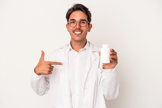 Jonge apotheker gemengd ras man met pillen geïsoleerd op een witte achtergrond persoon met de hand wijzend naar een shirt kopie ruimte, trots en zelfverzekerd?
