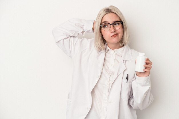 Jonge apotheker vrouw met pillen geïsoleerd op een witte achtergrond achterhoofd aan te raken, denken en een keuze maken.