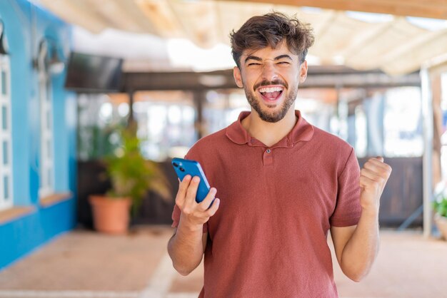 Foto jonge arabische knappe man buitenshuis met behulp van mobiele telefoon terwijl hij duimen omhoog doet