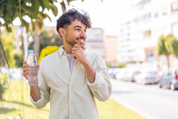 Jonge Arabische knappe man met een fles water buitenshuis die een idee denkt en naar de kant kijkt