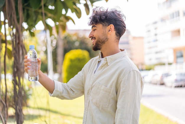 Jonge Arabische knappe man met een fles water in de buitenlucht met een gelukkige uitdrukking