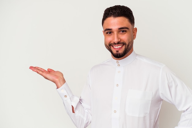 Jonge Arabische man die typische Arabische kleding draagt die op witte achtergrond wordt geïsoleerd die een exemplaarruimte op een palm toont en een andere hand op taille houdt.