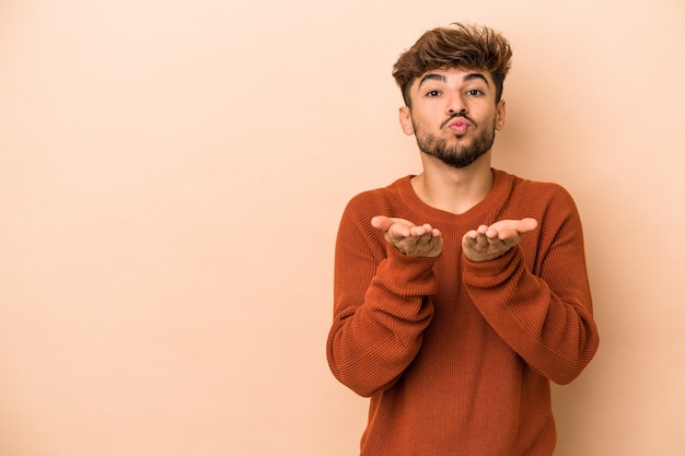 Jonge Arabische man geïsoleerd op beige achtergrond die lippen vouwt en handpalmen vasthoudt om luchtkus te sturen.