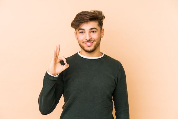 Jonge arabische man geïsoleerd op een beige muur vrolijk en zelfverzekerd tonend ok gebaar.