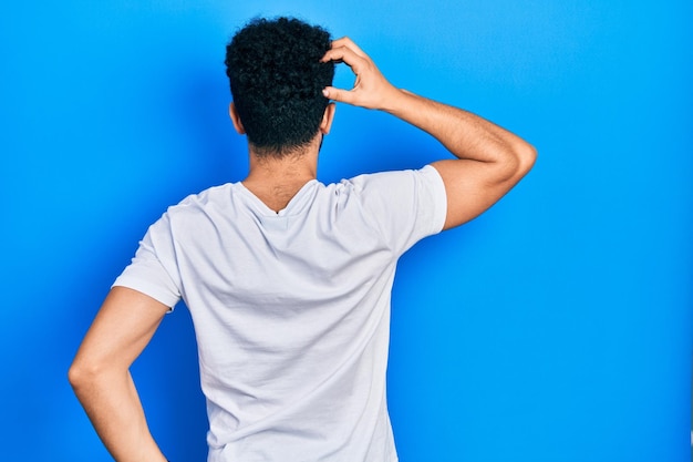 Jonge arabische man met baard die een casual wit t-shirt draagt en achterwaarts nadenkt over twijfel met de hand op het hoofd