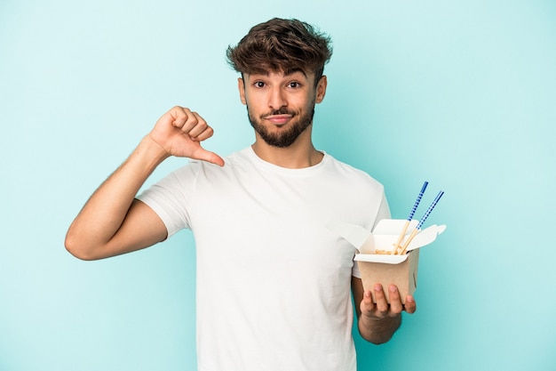 Jonge arabische man met een take-away noedels geïsoleerd op blauwe achtergrond voelt zich trots en zelfverzekerd, voorbeeld om te volgen.