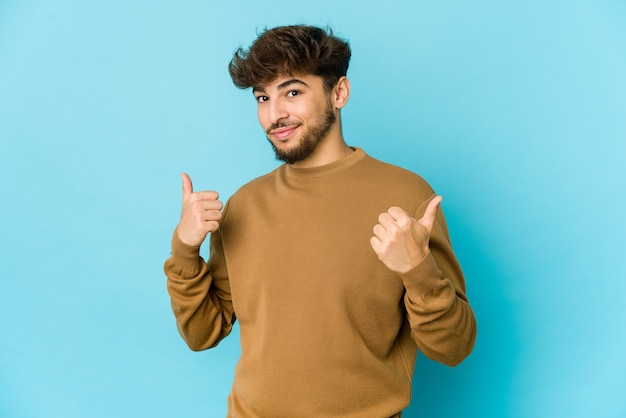 Jonge Arabische man op blauw die beide duimen opheft, glimlachend en zelfverzekerd.