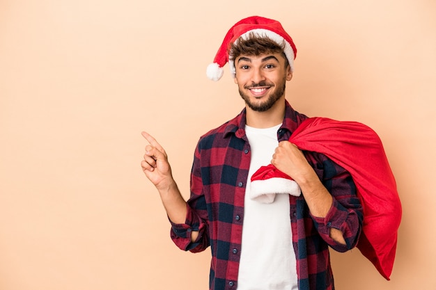 Jonge arabische man vermomd als de kerstman geïsoleerd op een beige achtergrond glimlachend en opzij wijzend, iets tonend op de lege ruimte.
