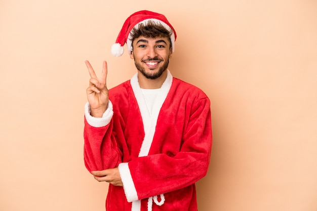 Jonge arabische man vermomd als de kerstman geïsoleerd op een beige achtergrond met nummer twee met vingers.