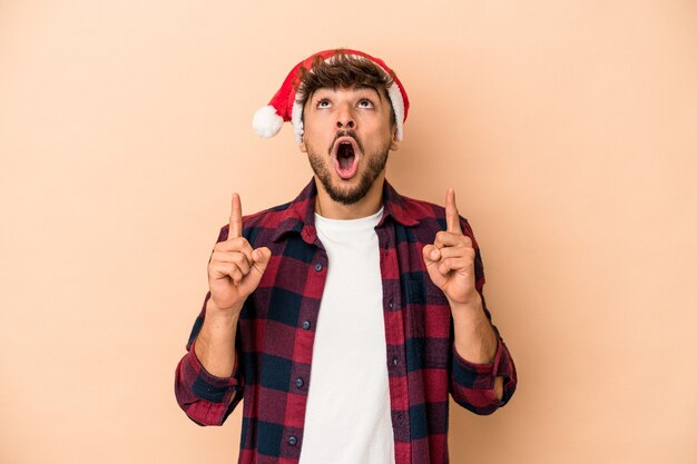 Jonge Arabische man viert Kerstmis geïsoleerd op beige achtergrond wijzend ondersteboven met geopende mond.