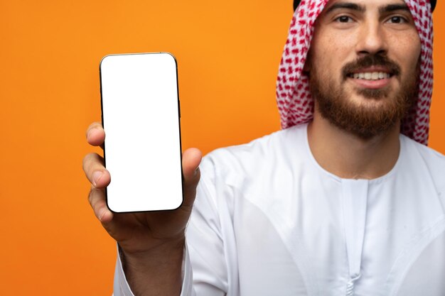 Jonge Arabische moslim man in traditionele kleding met een mobiele telefoon geïsoleerd op een oranje achtergrond