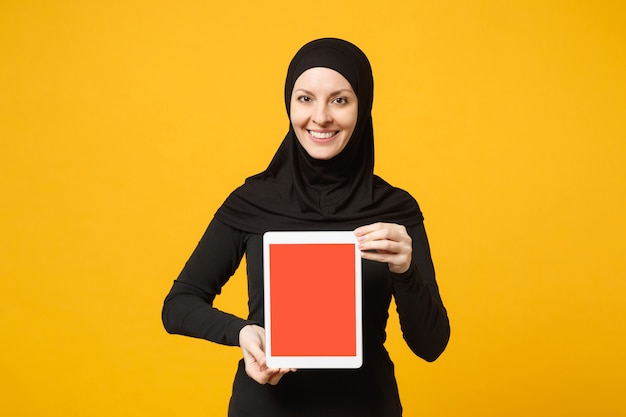 Jonge Arabische moslim werknemer vrouw in hijab zwarte kleren houden en werken tablet pc-computer geïsoleerd op gele muur portret. Mensen religieuze levensstijl concept.