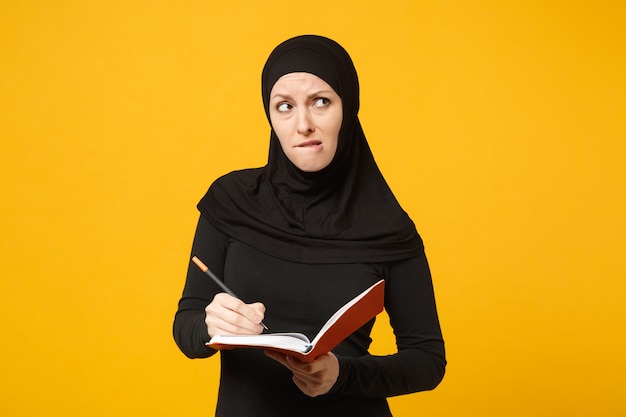 Jonge arabische moslimvrouw in hijab zwarte kleding houdt notitieboekje, schrijft, bereidt zich voor op examen geïsoleerd op geel muurportret. Mensen religieuze levensstijl concept. .