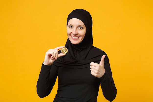 Jonge Arabische moslimvrouw in hijab zwarte kleren houden in de hand met bitcoin munt valuta geïsoleerd op gele muur portret. Mensen religieuze levensstijl concept.