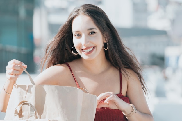 Jonge arabische poc-vrouw blij met haar nieuwe aankoop die naar de camera kijkt en glimlacht terwijl ze een boodschappentas opent, trendy moderne kleding draagt. Uitrusten na een dagje winkelen in het winkelcentrum, jeans en top.