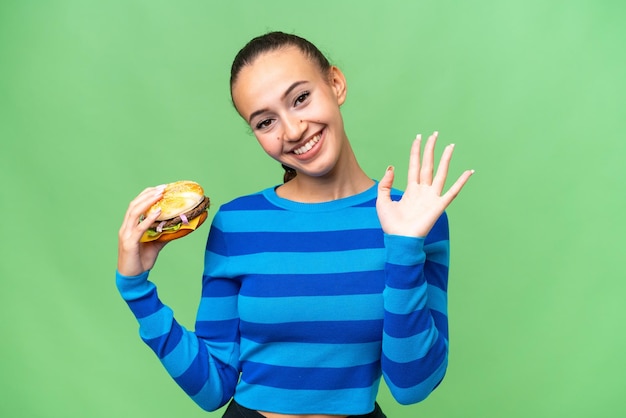 Jonge Arabische vrouw die een hamburger over een geïsoleerde achtergrond houdt en met de hand groet met een gelukkige uitdrukking