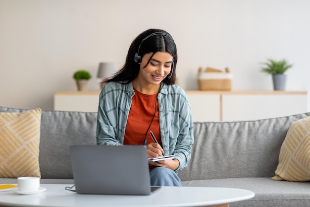 Jonge Arabische vrouw in koptelefoon met laptop die thuis werkt of studeert en aantekeningen maakt tijdens zakelijke bijeenkomst