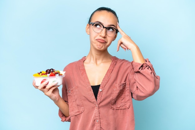 Jonge Arabische vrouw met een schaal fruit geïsoleerd op blauwe achtergrond met twijfels en denken