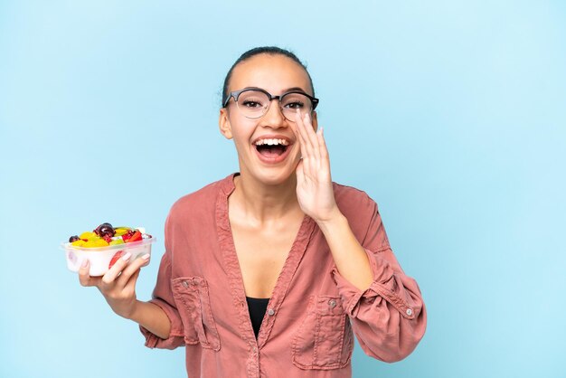 Jonge Arabische vrouw met een schaal fruit geïsoleerd op blauwe achtergrond schreeuwend met wijd open mond