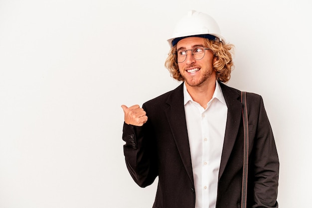 Foto jonge architect blanke man met helm geïsoleerd op een witte achtergrond wijst met duimvinger weg, lachend en zorgeloos.
