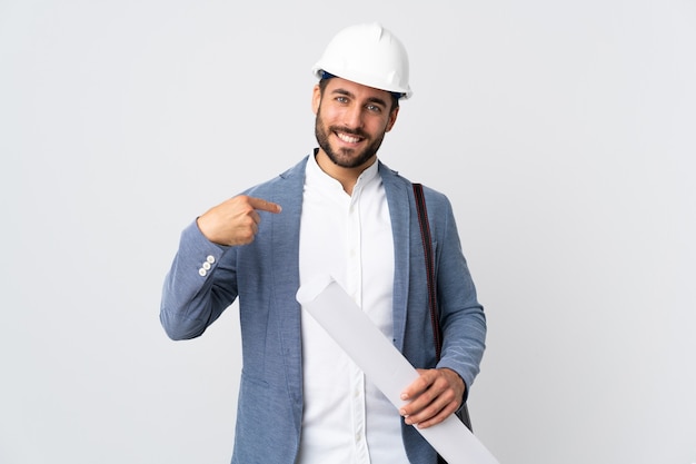 Jonge architect man met helm en blauwdrukken geïsoleerd op een witte muur trots en zelfvoldaan te houden