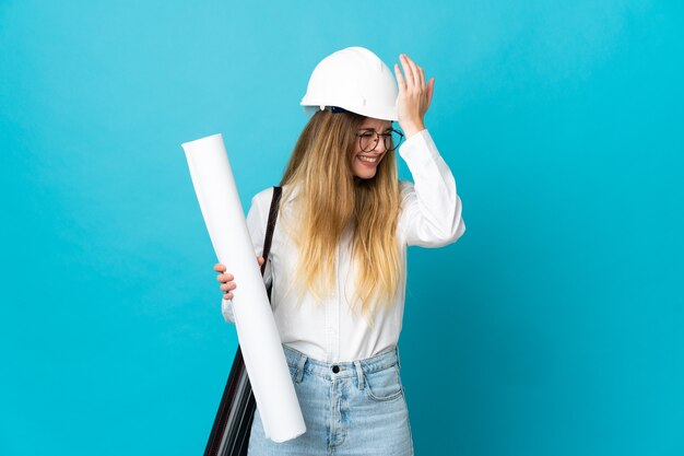 Jonge architectenvrouw met helm en blauwdrukken vasthouden die op blauwe muur worden geïsoleerd, heeft iets gerealiseerd en de oplossing beoogd