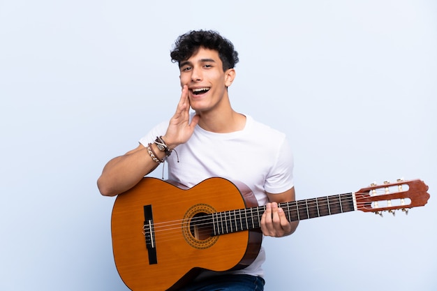 Jonge Argentijnse man met gitaar over geïsoleerde blauwe muur schreeuwen met wijd open mond