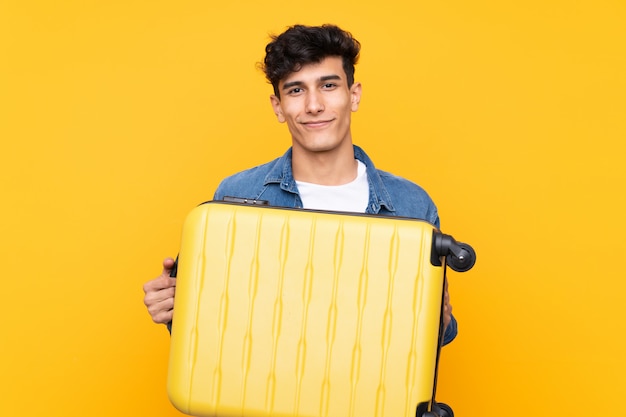 Jonge Argentijnse man over geïsoleerde gele achtergrond in vakantie met reiskoffer