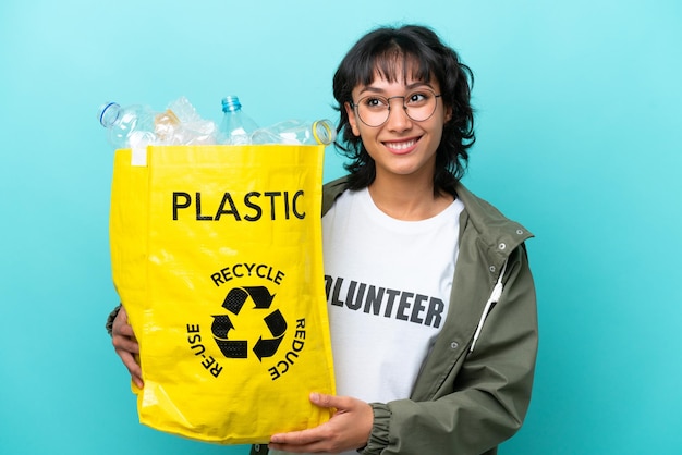 Jonge Argentijnse vrouw die een zak vol plastic flessen vasthoudt om te recyclen, geïsoleerd op een blauwe achtergrond en een idee denkt terwijl ze omhoog kijkt