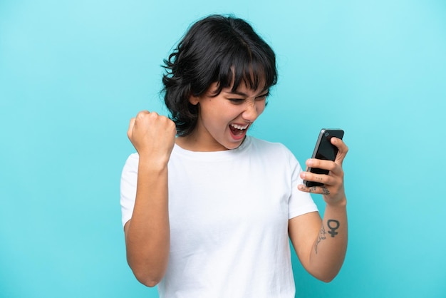 Jonge argentijnse vrouw geïsoleerd op blauwe achtergrond met behulp van mobiele telefoon en overwinningsgebaar doen