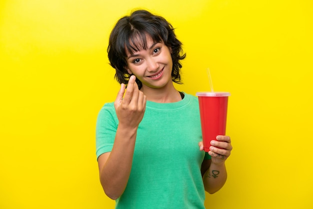 Jonge Argentijnse vrouw met een frisdrank geïsoleerd op een gele achtergrond die uitnodigt om met de hand te komen Blij dat je bent gekomen