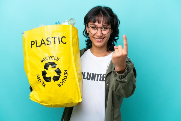 Jonge Argentijnse vrouw met een zak vol plastic flessen om te recyclen geïsoleerd op blauwe achtergrond die een komend gebaar doet