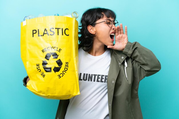 Jonge Argentijnse vrouw met een zak vol plastic flessen om te recyclen, geïsoleerd op een blauwe achtergrond, schreeuwend met de mond wijd open naar de zijkant