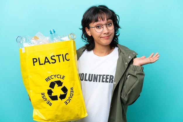 Jonge argentijnse vrouw met een zak vol plastic flessen om te recyclen, geïsoleerd op een blauwe achtergrond, twijfelt terwijl ze haar handen opsteekt