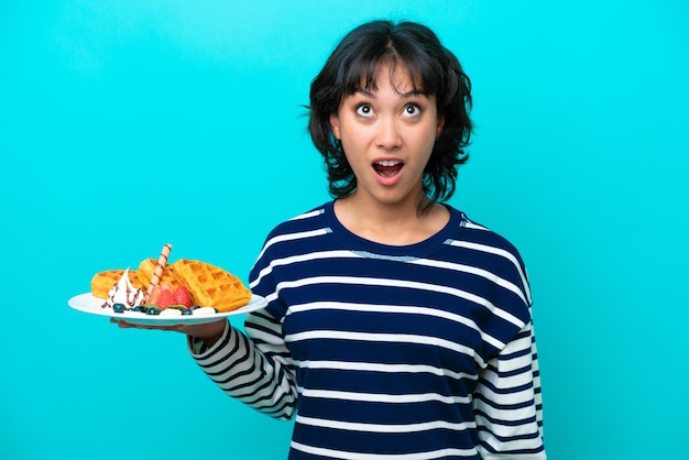 Jonge Argentijnse vrouw met wafels geïsoleerd op blauwe achtergrond opzoeken en met verbaasde uitdrukking