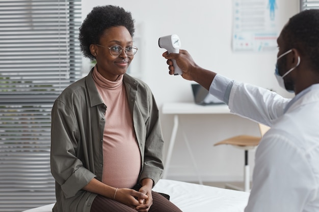 Jonge arts die de temperatuur van de zwangere vrouw onderzoekt tijdens haar bezoek aan het ziekenhuis