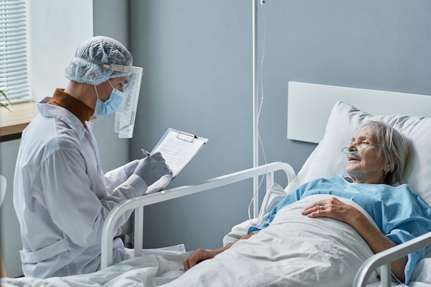 Jonge arts in beschermende kleding die een recept op een medische kaart schrijft tijdens een bezoek aan een oudere vrouw op de ziekenhuisafdeling